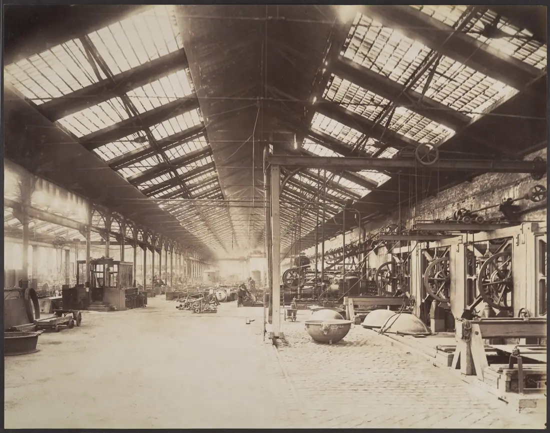 Fotografia antiga, em tons de sépia, do interior de uma grande oficina de ferro: um telhado de vidro e vigas metálicas se estende até o fundo; no alto, à direita, eixos horizontais com polias de onde descem correias em direção a máquinas de grandes rodas; no chão de pedra, bacias metálicas redondas e vagonetes.